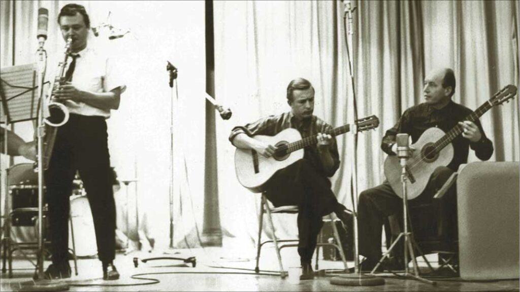 a male saxophonist stands while playing next to a pair of seated men playing their guitars