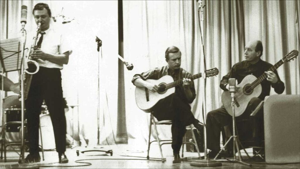 A saxophonist and two guitar player perfroming on a small stage with microphones.